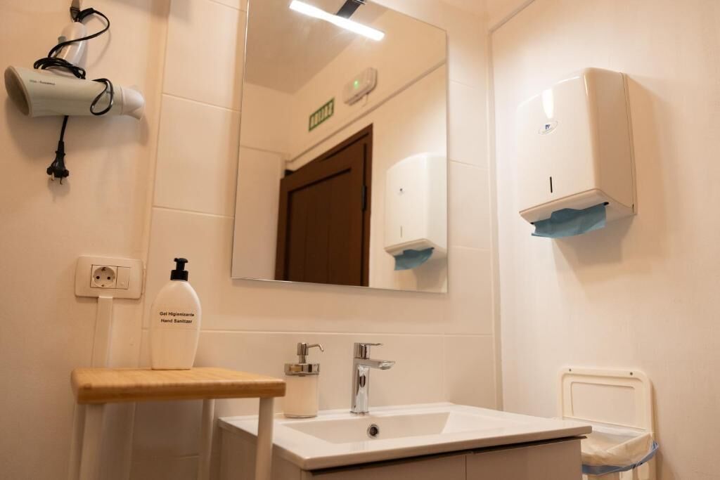 Baño con lavamanos, dispensador de gel antibacterial, espejo, secador de pelo y secador de manos.