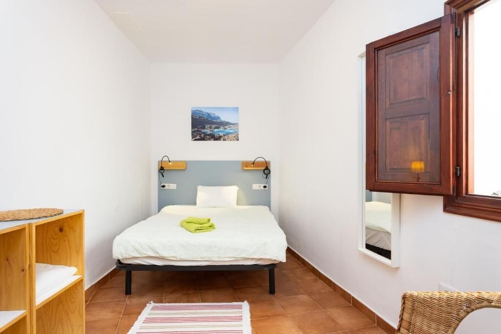 Una habitación pequeña y estrecha con una cama, un estante de madera, una silla de mimbre y una ventana con vistas.