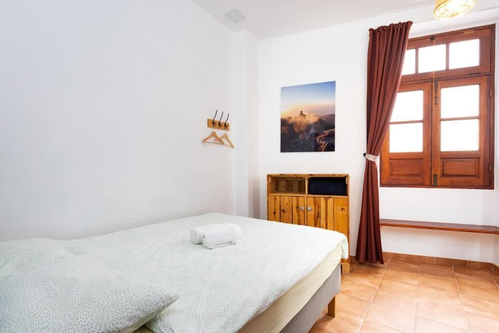 Uma cama de solteiro com cobertor branco, um armário de madeira, uma janela com cortinas e um quadro fixado na parede.