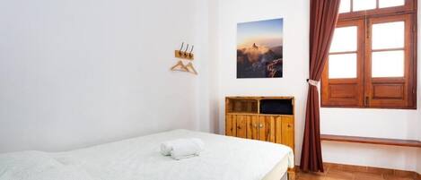 Uma cama de solteiro com cobertor branco, um armário de madeira, uma janela com cortinas e um quadro fixado na parede.