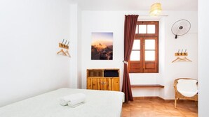 Une pièce comprenant un lit, une armoire en bois, une fenêtre avec des rideaux, un ventilateur fixé au mur et un tableau accroché au mur.