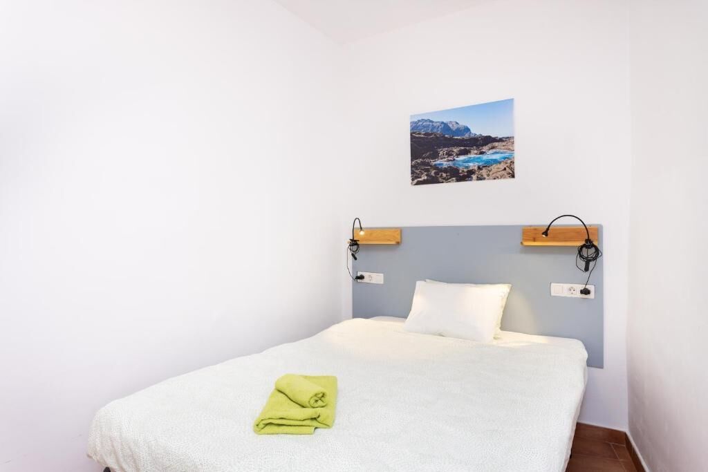A single bed with a white mattress and a green towel, a wooden headboard, a wall-mounted picture of a coastal landscape, and a small wooden shelf.