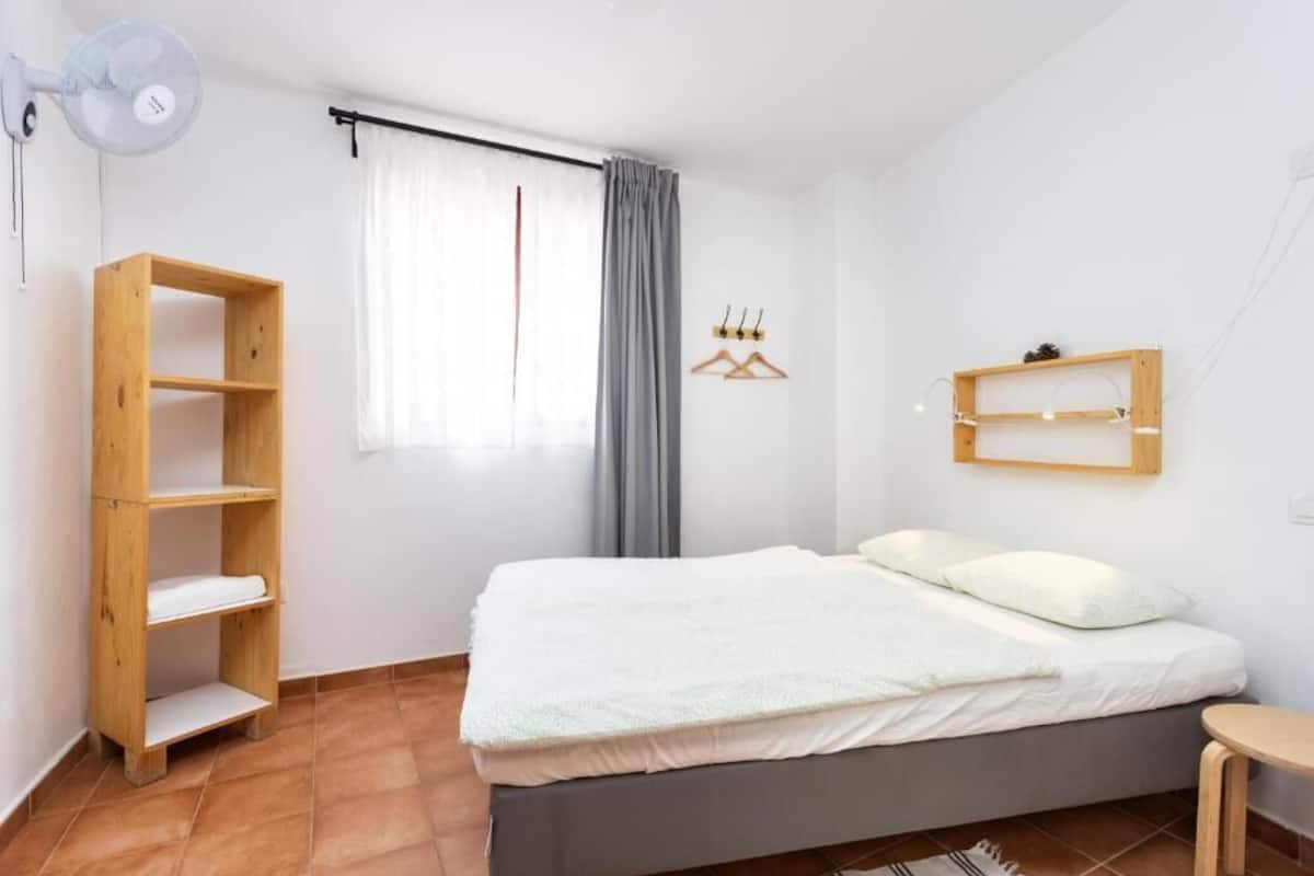 Une chambre à coucher avec un lit, une étagère en bois, un ventilateur, une fenêtre avec des rideaux et une étagère fixée au mur.