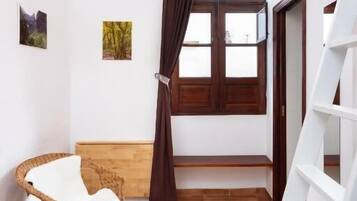 A room with a wicker chair, a white chair, a white shelf, a window with brown curtains, and a white ladder.