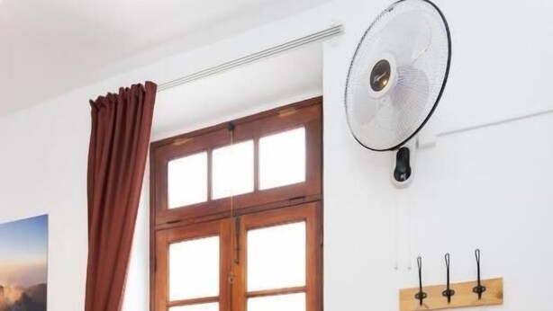 Une pièce comprenant un banc en bois, une chaise en osier, une fenêtre avec des rideaux, un ventilateur fixé au mur et une armoire en bois.