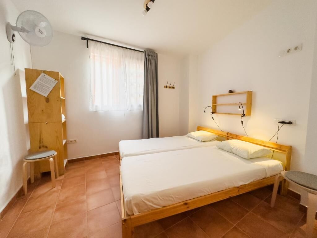 Et soveværelse med to senge, et skrivebord, en stol, en ventilator og et vindue med gardiner.