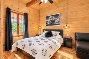 Un dormitori de cabana de fusta amb un llit, dues làmpades de nit, una cadira de cuir negre i una fotografia emmarcada a la paret.