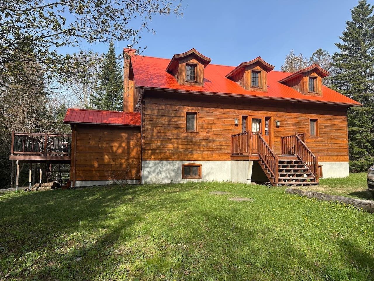 A two-story wooden cabin with a red roof, a wooden deck, and a staircase leading to the upper floor.