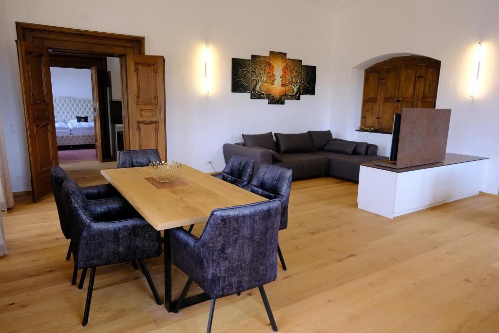Una sala de estar moderna con una mesa de comedor de madera, sillas tapizadas oscuras, un sofá gris y una obra de arte colgada en la pared.