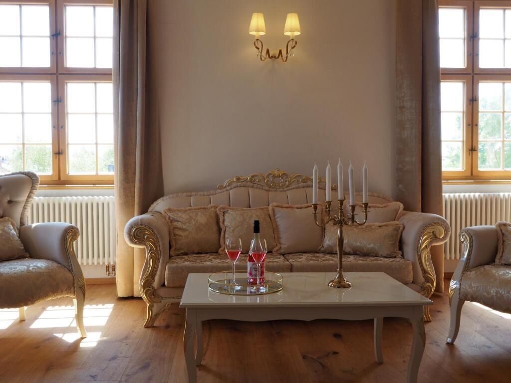 A room with a large window, a sofa, two armchairs, a table with a candelabra, and two lamps on the wall.