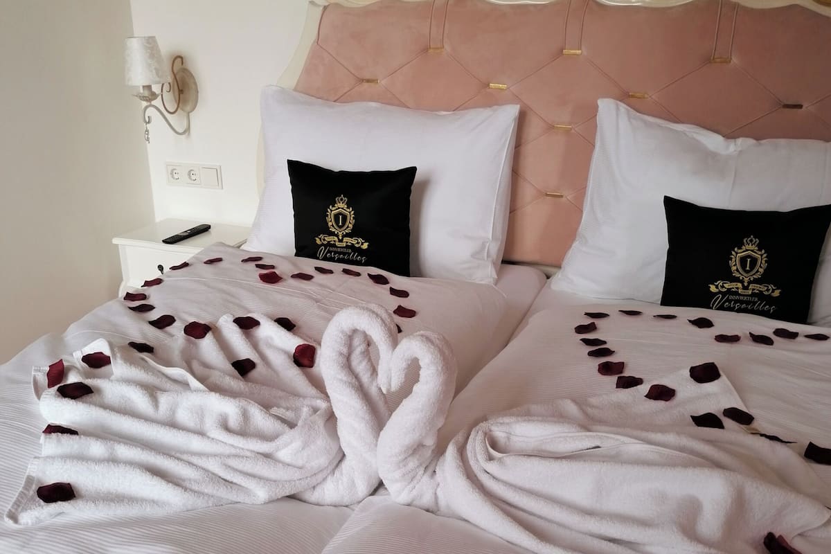 A neatly made bed with rose petals, a bedside table with a remote control, and a wall-mounted lamp.