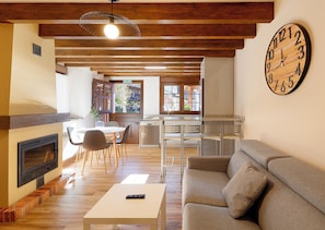 A spacious living area with a fireplace, a sofa, a dining table with chairs, a kitchen area, and a large wall clock.