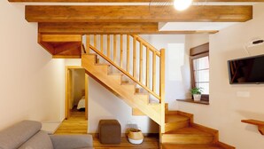 Eine Holztreppe führt zu einer oberen Etage, ein graues Sofa, ein kleiner Hocker, ein gewebter Korb und ein Fernseher an der Wand.