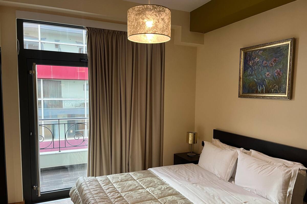 Habitación de hotel con cama, un cuadro, una lámpara, una puerta corrediza a un balcón y vistas de edificios.