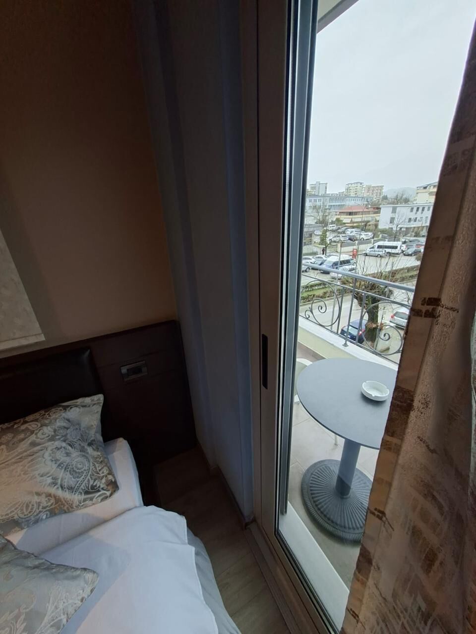 A hotel room with a bed, a window with curtains, and a view of a cityscape.