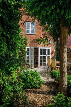 A brick house with a large window and a sliding glass door, surrounded by greenery and a patio area. - Historic house in Gammendorf on Fehmarn (Fehmarn)