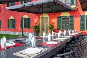 A dining table set with plates, glasses, and cutlery, surrounded by chairs, under a large umbrella, with a building featuring green shutters and a red wall in the background.