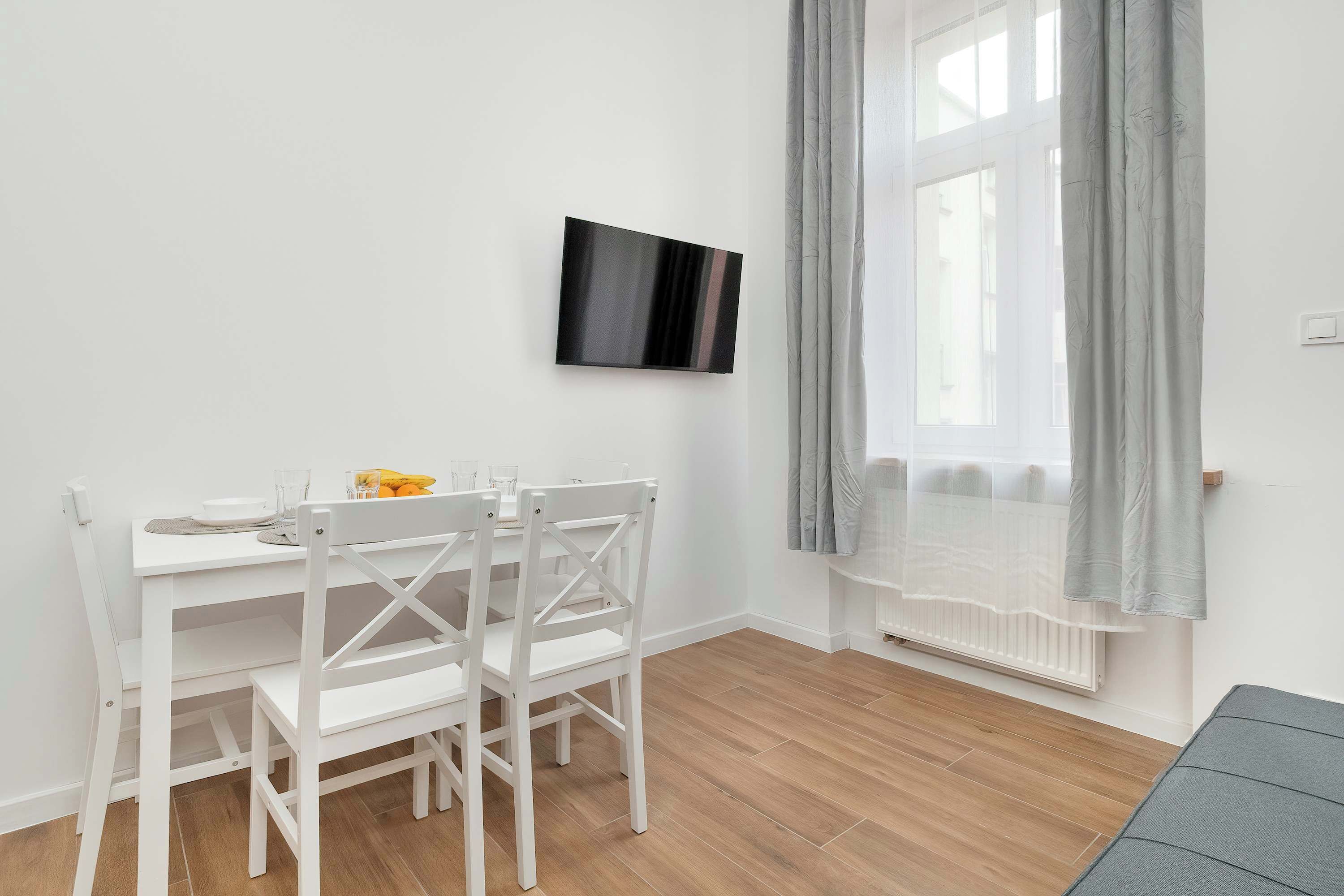 A white dining table with chairs, a flat-screen TV mounted on the wall, a window with grey curtains, and a radiator.