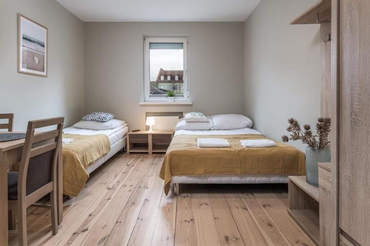 A room with two beds, a desk, a chair, a window, and a view of a building outside.