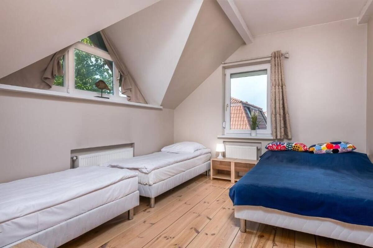 A room with two single beds, a window with curtains, a radiator, and a small table with a lamp.
