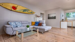 A modern living room with a grey sofa, a glass coffee table, and a surfboard decoration on the wall.