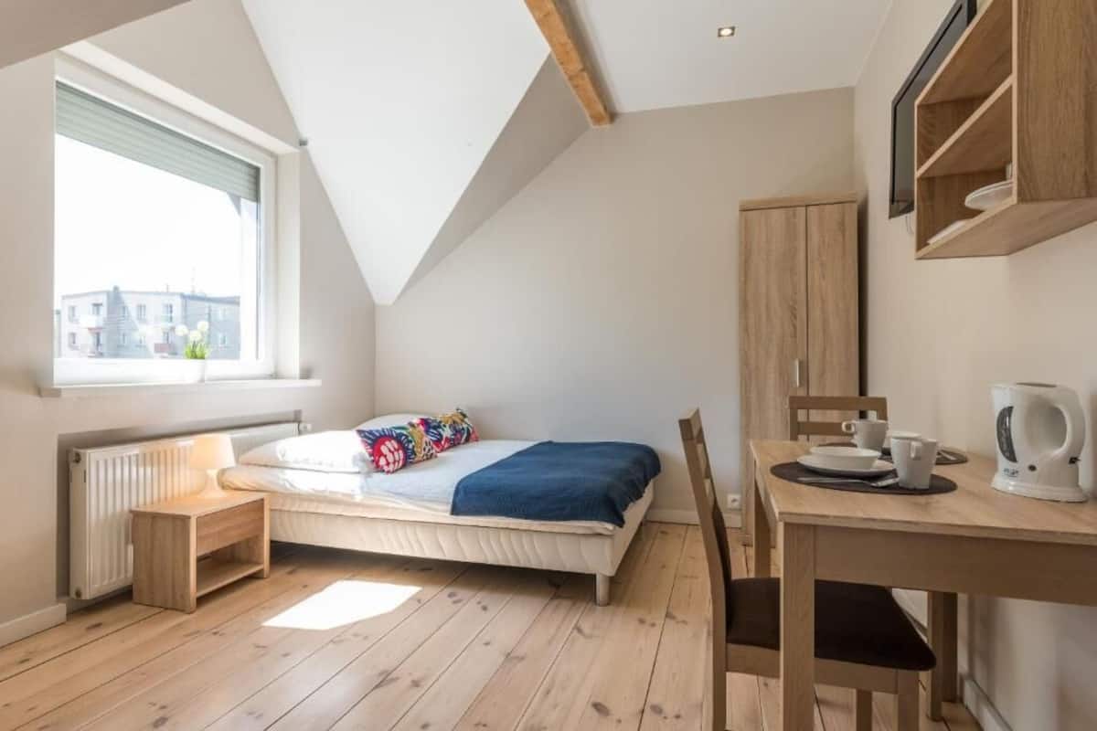 A bedroom with a bed, a window, a radiator, a wooden table with a kettle, and a wooden cabinet.