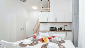 A modern kitchen with white cabinets, a wooden staircase, and a dining area with white chairs and a table set for two. - Functional Studio with Mezzanine | Łódź Center (Łódź)