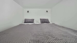 A small, single-bed room with a textured grey bedspread, two pillows, and a white wall with two wall-mounted lights. - Functional Studio with Mezzanine | Łódź Center (Łódź)