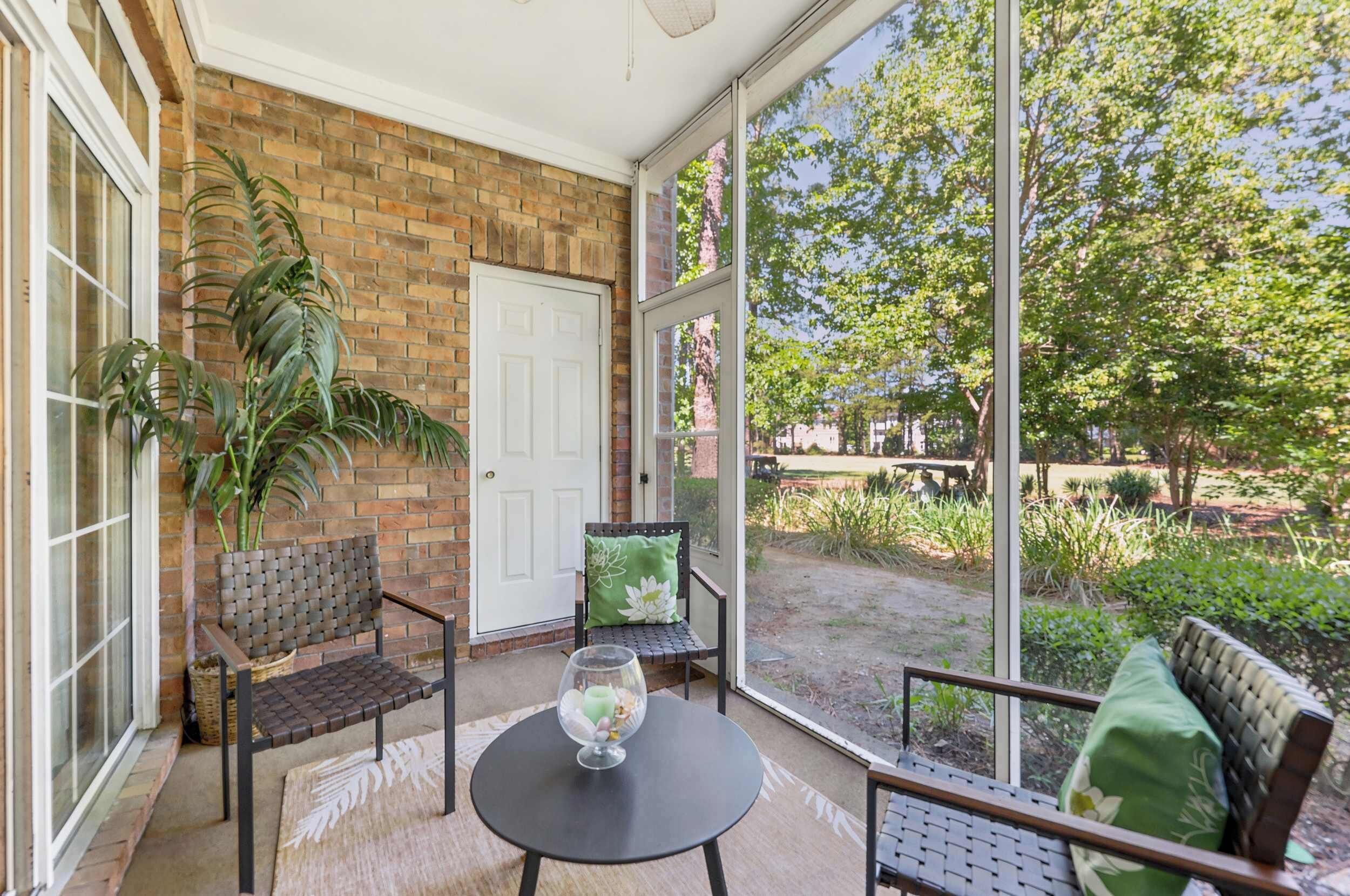 A covered patio with a brick wall, a white door, two chairs, a table with a glass object, and a view of a garden and trees.
