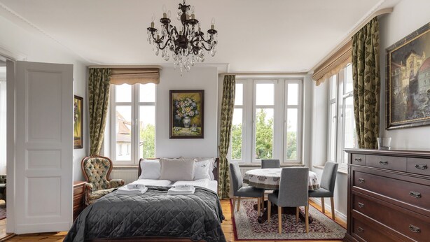 A bedroom with a bed, a chandelier, a dining table with chairs, a dresser, and a painting on the wall. - Grand Belle Sopot - Near the beach (Sopot)