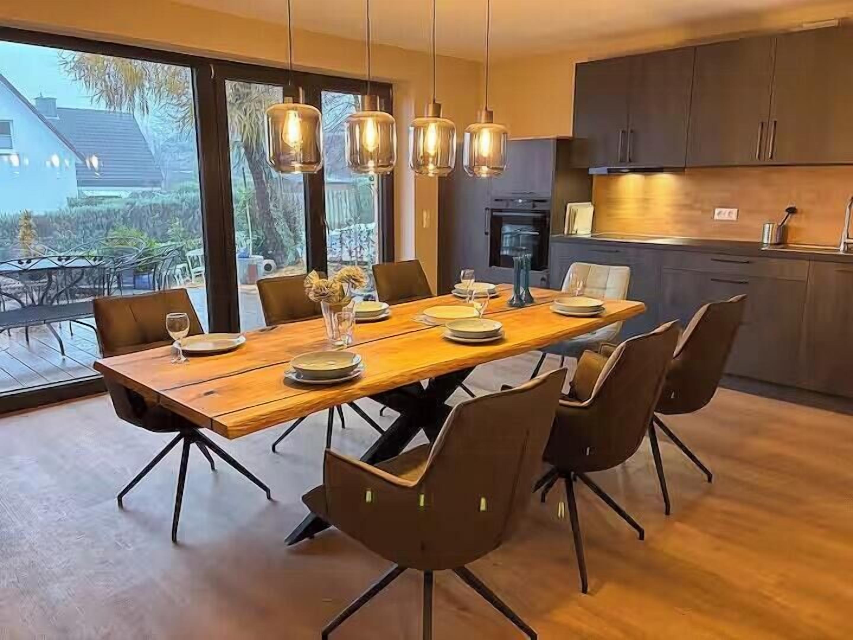 Uma área de jantar moderna com mesa e cadeiras de madeira, luminárias pendentes e vista para o exterior.