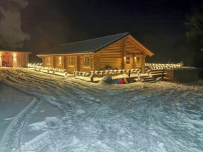 A log cabin with a porch, illuminated by string lights, surrounded by snow. - Round timber house in forest near Mariager (Hadsund)