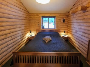 A log cabin bedroom with a bed, two bedside tables, a window, and a wall-mounted picture. - Round timber house in forest near Mariager (Hadsund)
