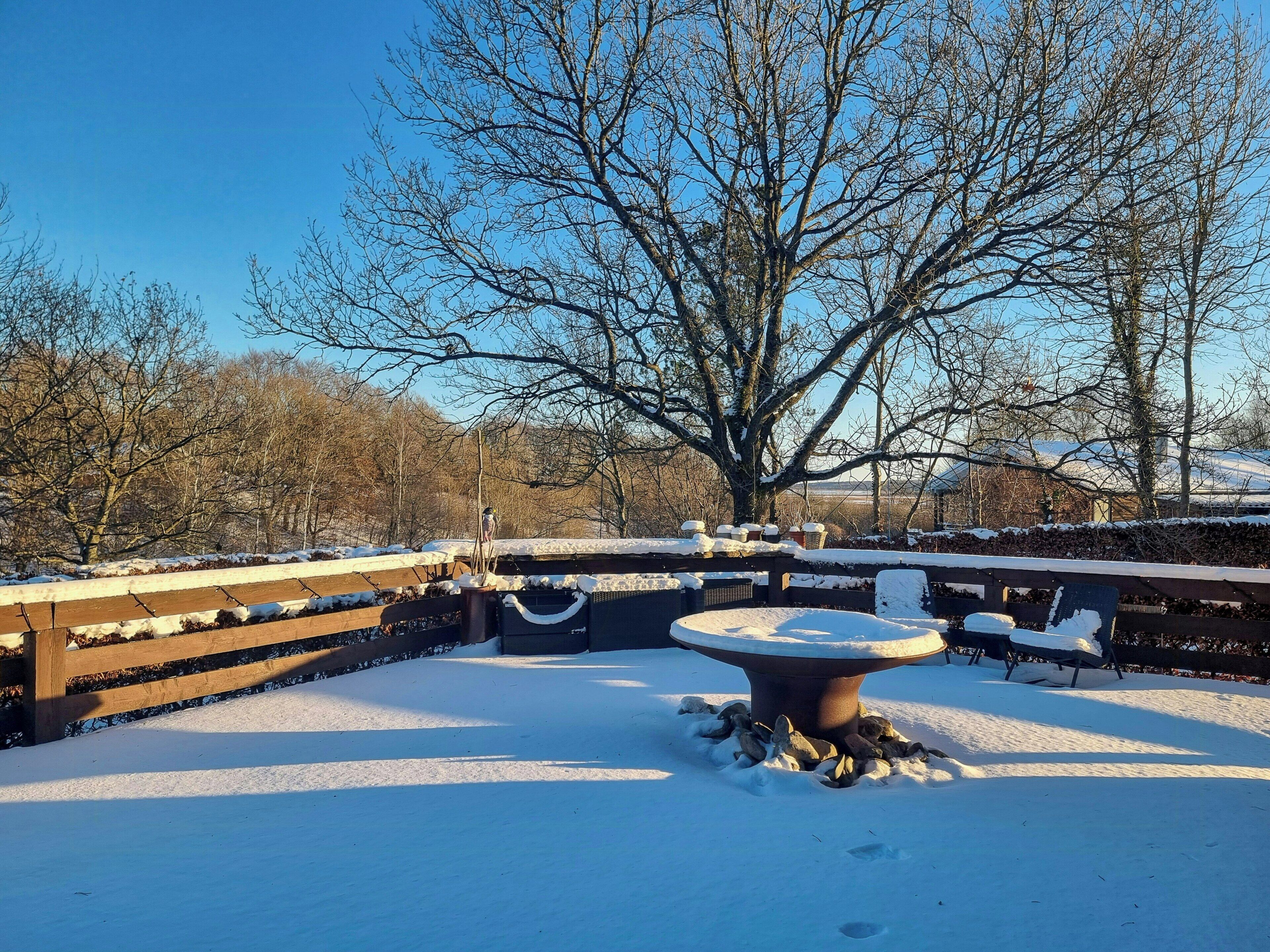 A snow-covered outdoor area with a stone table, benches, and a wooden fence.