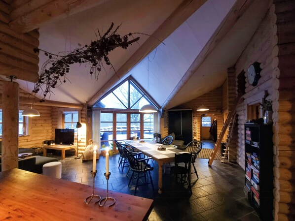 A log cabin interior with a dining area, a fireplace, and a large window overlooking a snowy landscape. - Round timber house in forest near Mariager (Hadsund)