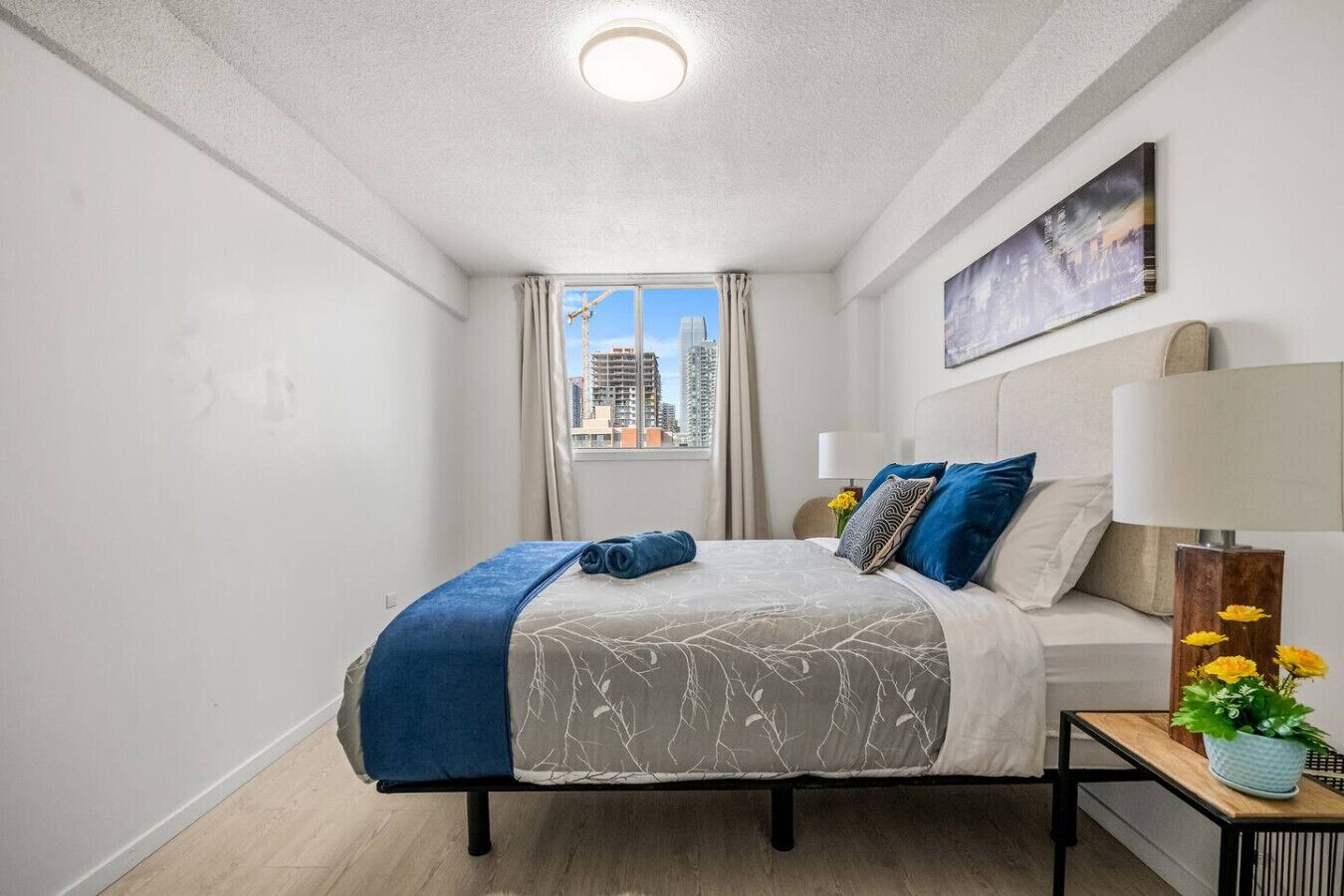 A hotel room with a bed, a nightstand, a lamp, a window with a city view, and a painting on the wall.