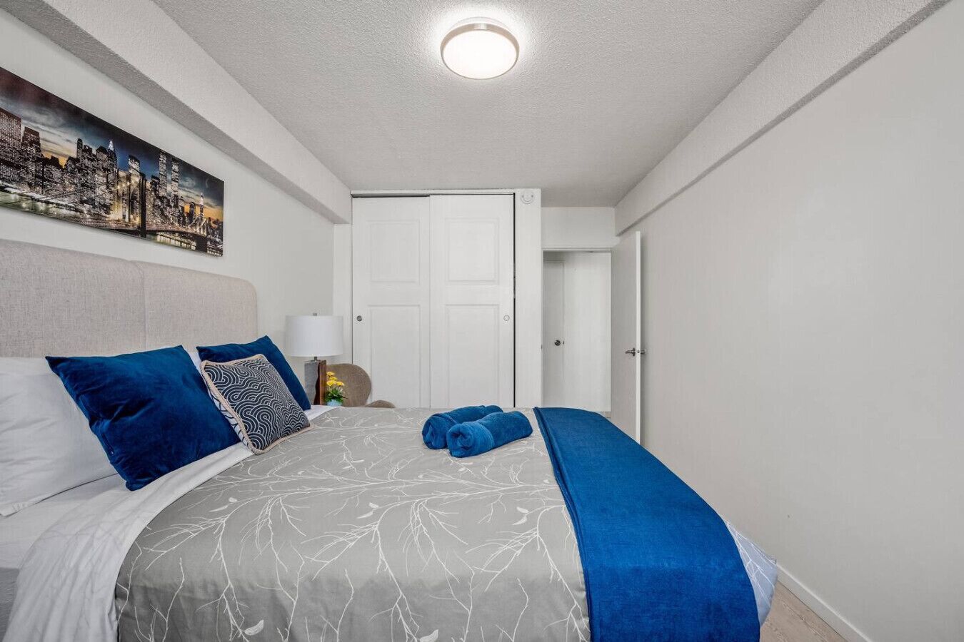 A neatly arranged bedroom with a bed, pillows, a bedside table, a lamp, a door, and a cityscape painting on the wall.