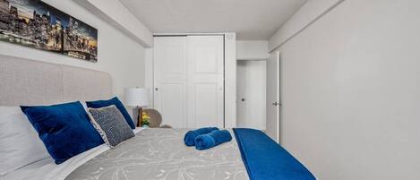 A neatly arranged bedroom with a bed, pillows, a bedside table, a lamp, a door, and a cityscape painting on the wall.