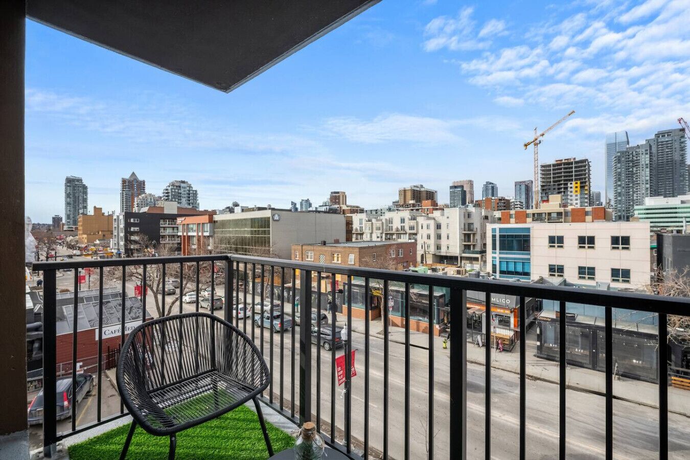A balcony with a chair, a view of a cityscape with buildings and construction cranes, and a clear sky.