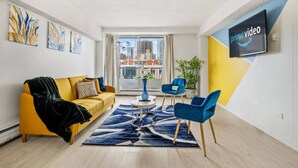 Un salon moderne avec un canapé jaune, des fauteuils bleus, une table basse et une grande fenêtre donnant sur la ville.