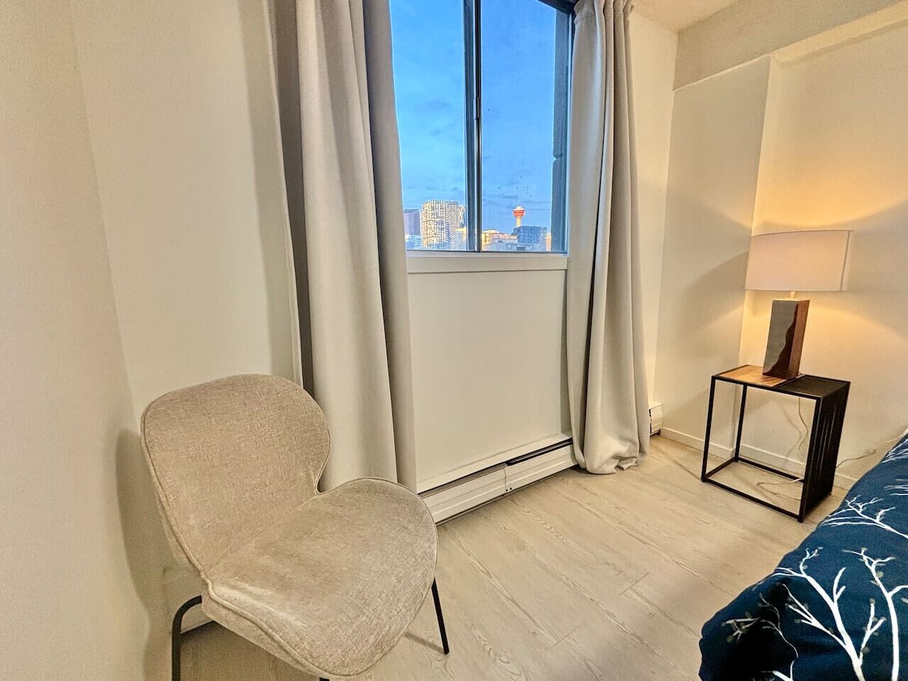 A room with a chair, a bed with a blue patterned bedspread, a nightstand with a lamp, and a view of a cityscape through a window with curtains.