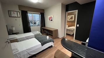 Quarto de hotel com duas camas, uma escrivaninha, uma cadeira, uma janela com vista para a cidade e um banheiro visível por uma porta aberta.
