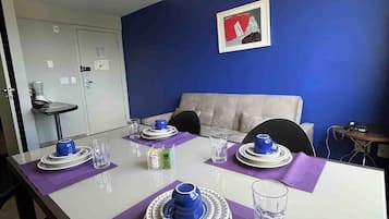 Uma área de refeições com uma mesa branca posta para quatro pessoas, jogos americanos roxos e louças azuis e brancas.