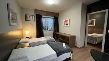 Quarto de hotel com duas camas, uma escrivaninha, uma cadeira, uma janela com vista para a cidade e uma porta que dá para outro quarto.