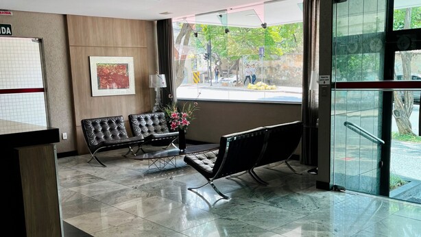 A lobby with a reception desk, two leather chairs, a vase with flowers, and a large window offering a view of the street. - LK Barro Preto 10 (Belo Horizonte)