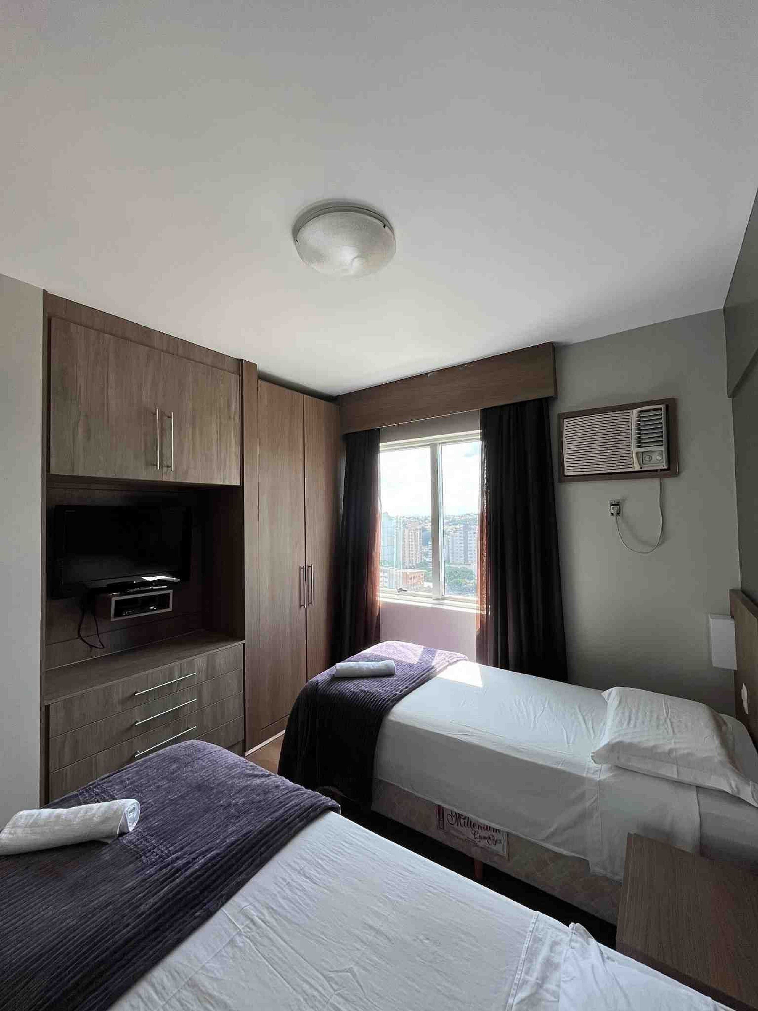 Quarto de hotel com duas camas, uma televisão sobre um móvel, janela com cortinas e vista para a cidade.