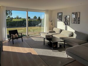 Un salon moderne avec un canapé, une table basse et un fauteuil. De grandes fenêtres sont ornées de rideaux, et des œuvres d’art décorent les murs.