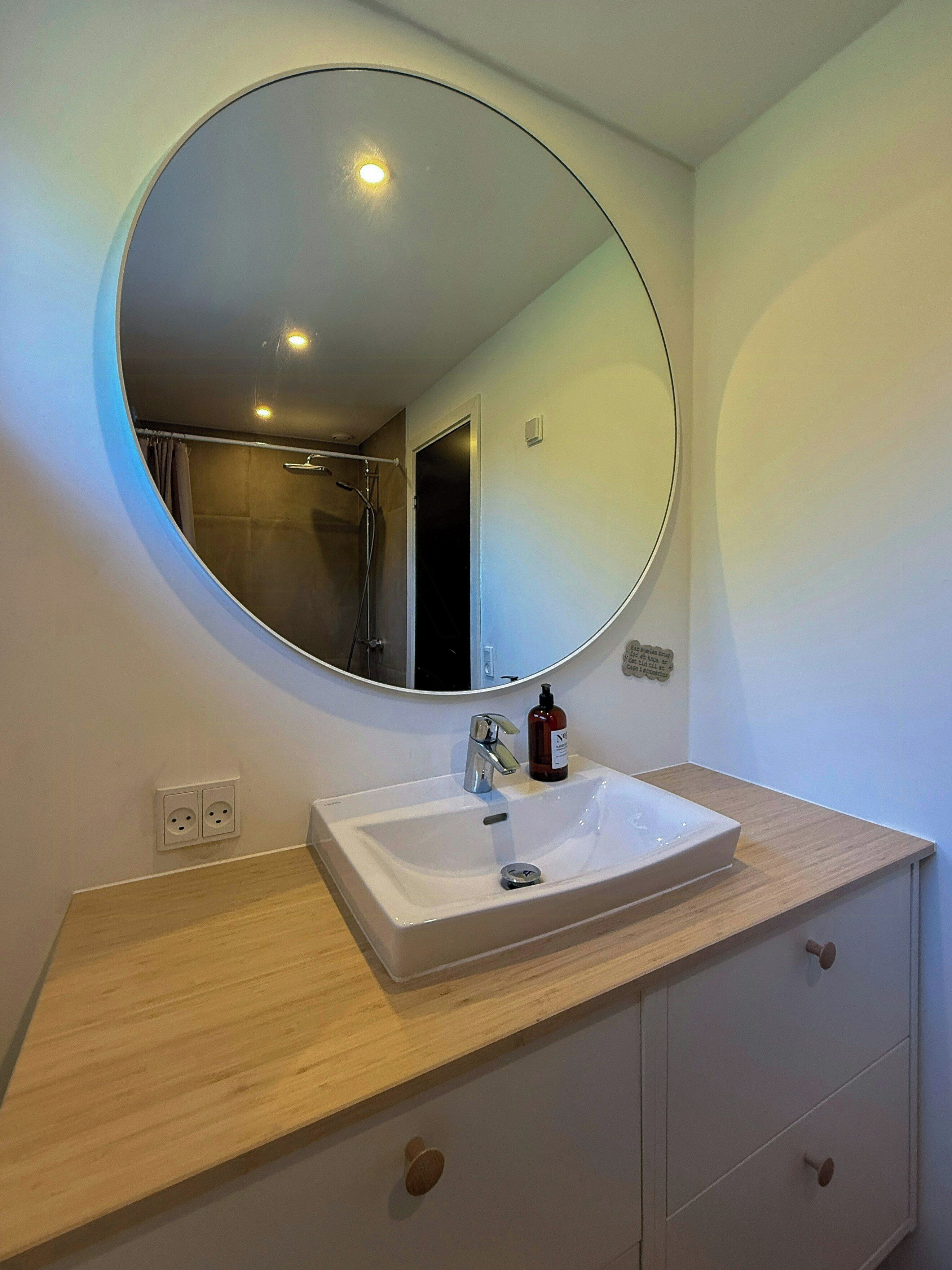 Un baño con un espejo redondo, un lavabo blanco, una encimera de madera y un mueble con cajones.