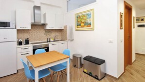 Une cuisine compacte avec des meubles blancs, un four à micro-ondes, une cuisinière et un évier. On y trouve une table à manger avec des chaises bleues et un tableau accroché au mur.