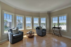 Una habitación amplia con ventanas grandes, dos sillones, una mesita redonda y una mesita auxiliar con una lámpara.